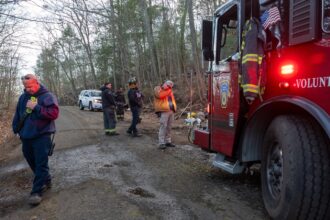 Firefighters mill around on a dirt road next to a red firetruck that says Volunteer on the side