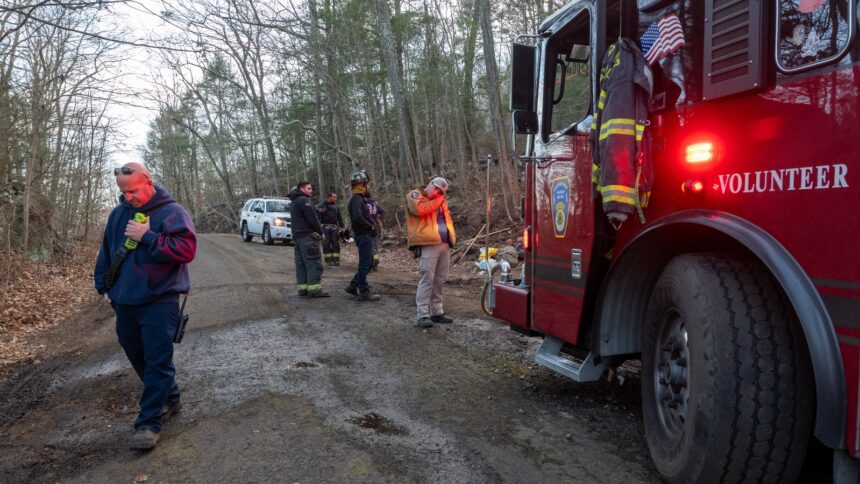 Firefighters mill around on a dirt road next to a red firetruck that says Volunteer on the side