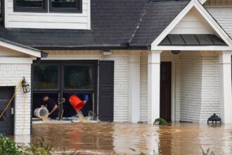 Two homeowners use buckets to bail water through the window of their flooded home.