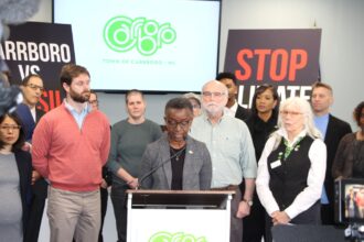 A woman in a gray jacket and black shirt stands at a podium with a group of people