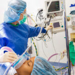 An anesthesiologist in blue scrubs and face mask applies anesthesia to a patient while looking at her monitors.