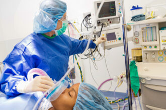 An anesthesiologist in blue scrubs and face mask applies anesthesia to a patient while looking at her monitors.