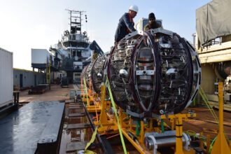 Workers on a ship prepare to launch a giant round piece of equipment off of a bright yellow ramp