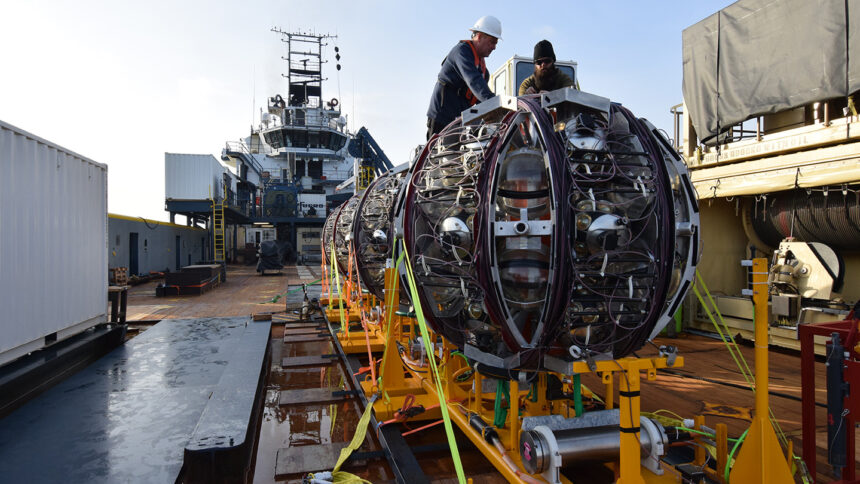 Workers on a ship prepare to launch a giant round piece of equipment off of a bright yellow ramp