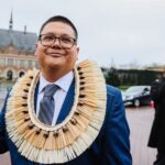 A man in a blue suit with shell necklace smiles in front of the Hague