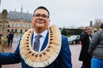 A man in a blue suit with shell necklace smiles in front of the Hague