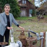 A woman wearing a blazer stands next to a water well with a farmhouse in the background