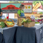 photo of two women selling fruits and vegetables from a stand under a tent at a farmers market, while a male customer looks over the merchandise