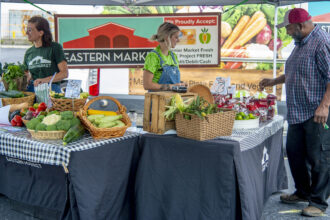 photo of two women selling fruits and vegetables from a stand under a tent at a farmers market, while a male customer looks over the merchandise