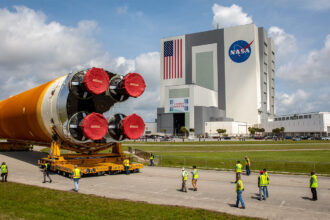 A huge rocket booster on its side, resting on two wheeled platforms, in front of a large blocky building. Workers in yellow vests walk nearby.