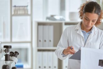 Female scientist reading notebook next to microscope in lab.