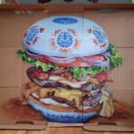 a painting of a juicy drippy burger with a blue and white porcelain style motif on the bun all on a cardboard box