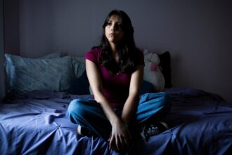 A teenage girl in a red tee shirt and jeans sits pensively on a bed