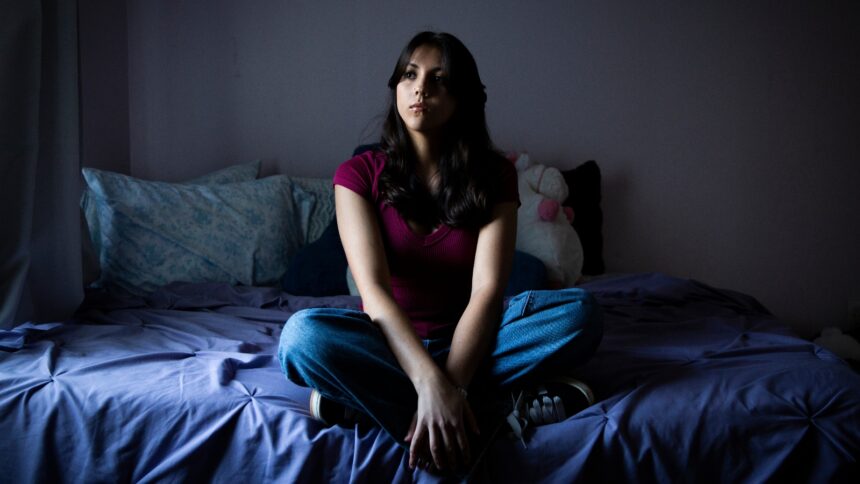 A teenage girl in a red tee shirt and jeans sits pensively on a bed