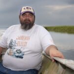 A bearded man in a white tee shirt and white baseball cap sits in a boat on a river