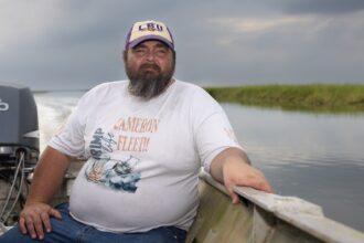 A bearded man in a white tee shirt and white baseball cap sits in a boat on a river