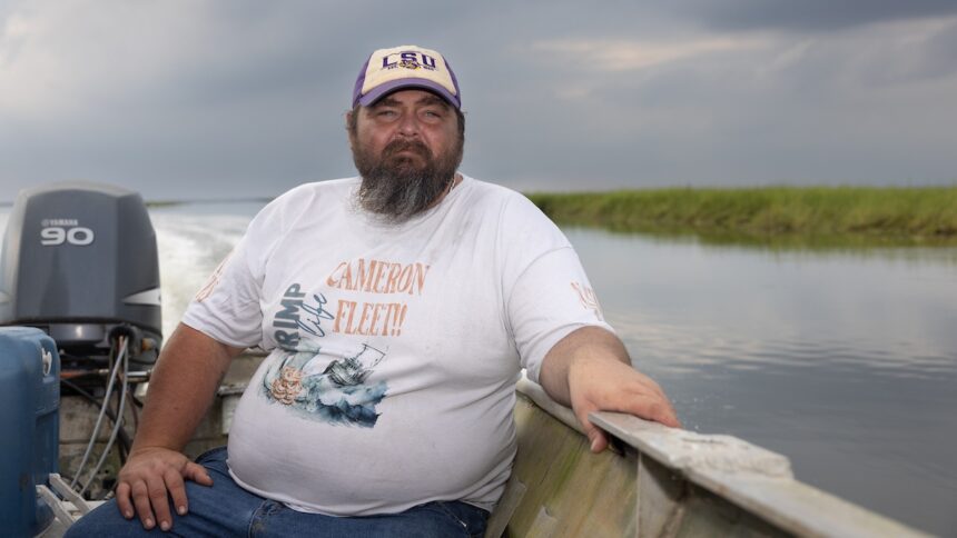 A bearded man in a white tee shirt and white baseball cap sits in a boat on a river