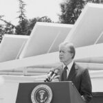 in a black and white photo, Jimmy Carter stands behind a podium with the presidential seal and in front of solar panels on the roof of the White House