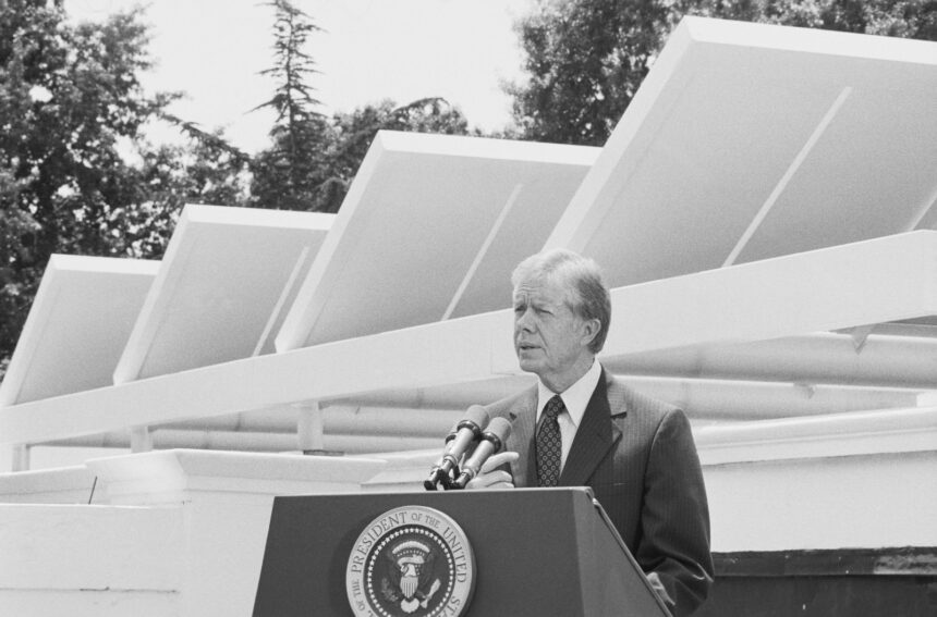 in a black and white photo, Jimmy Carter stands behind a podium with the presidential seal and in front of solar panels on the roof of the White House