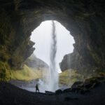 a figure stands in the opening of a cave, its silhouette accentuated against a waterfall