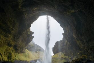 a figure stands in the opening of a cave, its silhouette accentuated against a waterfall