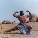 an indian woman in a a small checked bandeau top and blue and white checked skirt shows off her large muscles in a squat