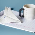 a mug and paper airplane sculpture that appear to made of lined paper atop white notepad