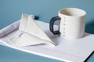 a mug and paper airplane sculpture that appear to made of lined paper atop white notepad