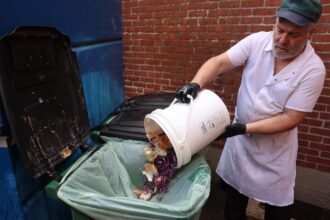 A restaurant worker empties out a bucket of food scraps into a trash can