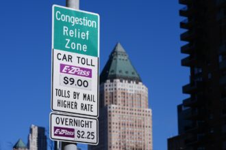 Close-up of a sign announcing congestion pricing tolls in New York City, with a blue sky and building in the background