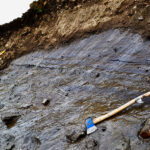 An exposed patch of gray, layered glacier ice is exposed in a brown, earthen headwall.