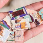 a hand holds up a collection of small, vibrant collages, each shaped like a stamp. they depict scenes with a building, water, architecture, and patterned designs