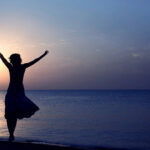 A happy woman with her arms up in the air at sunset on a beach.