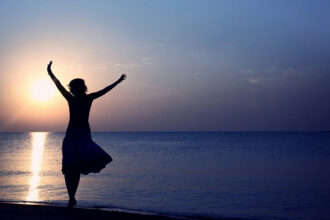 A happy woman with her arms up in the air at sunset on a beach.